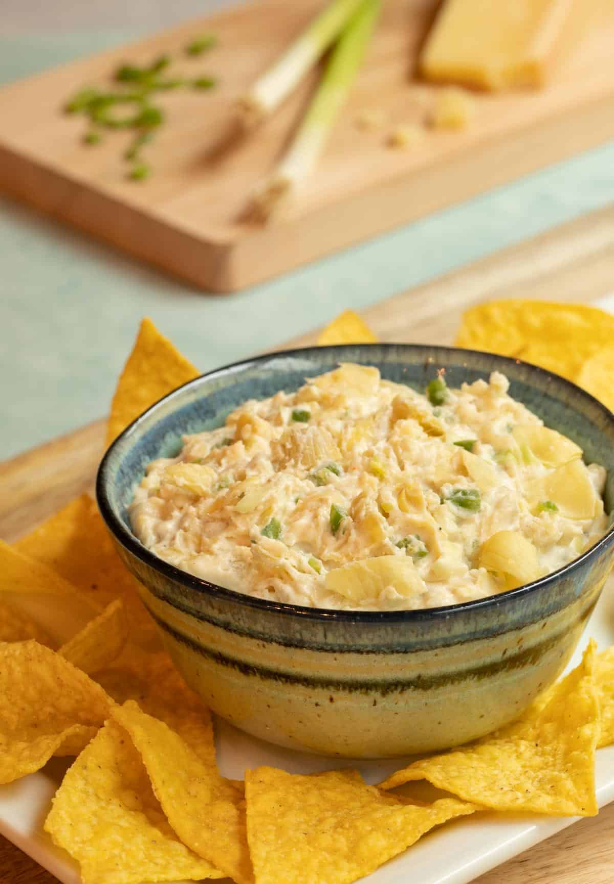 Blue bowl filled with Artichoke, Jalapeno and Cheese Dip, sitting on a white platter with tortilla chips