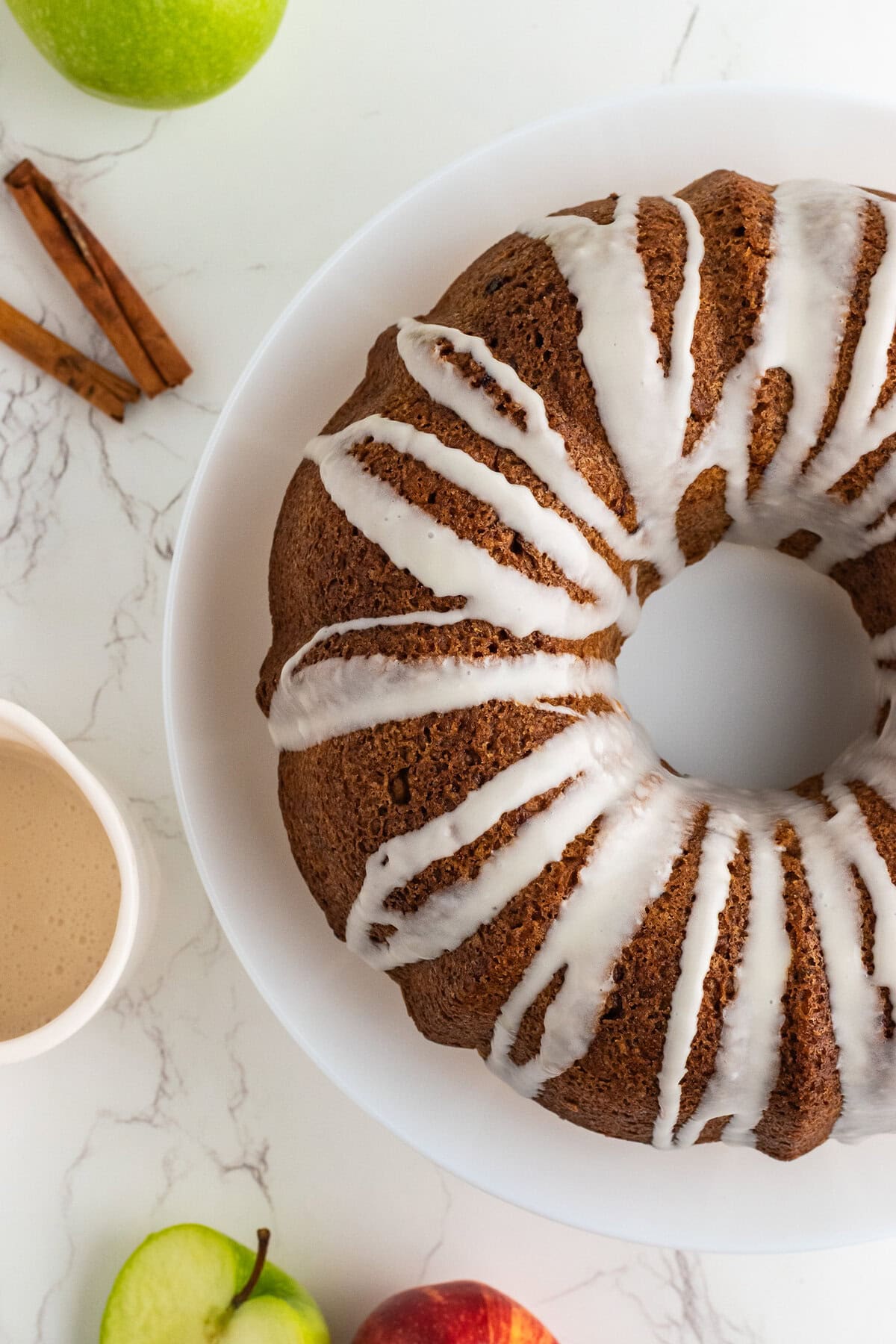 A glazed apple bundt cake on a table with half the cake showing and cinnamon sticks to the side.