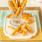 a glass holding cheese straws atop a white platter, with a few cheese straws in front and dipping sauce in the back
