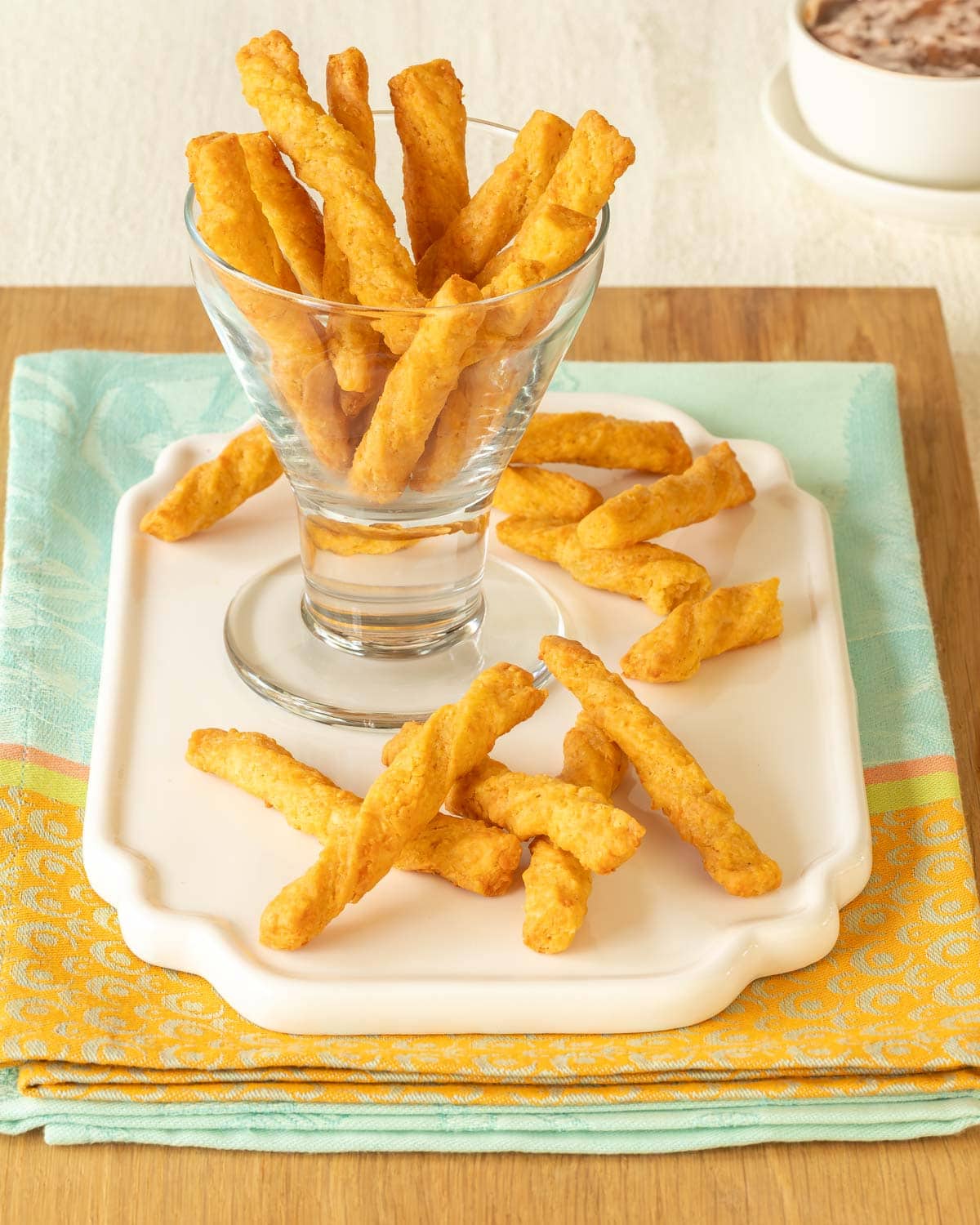 a glass holding cheese straws atop a white platter, with a few cheese straws in front and dipping sauce in the back
