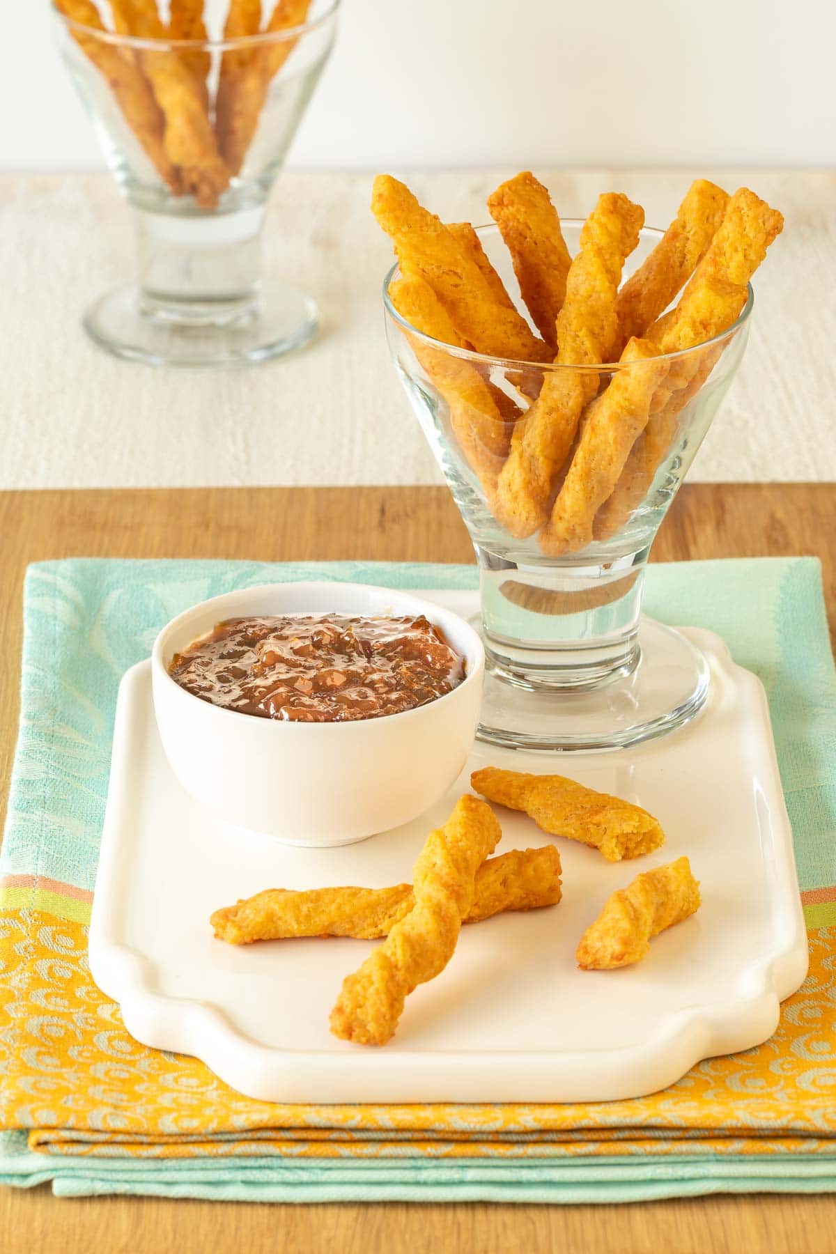 a glass holding cheese straws atop a white platter, with a few cheese straws in front and dipping sauce on the side