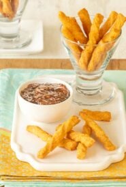 a glass holding cheese straws atop a white platter, with a few cheese straws in front and dipping sauce on the side