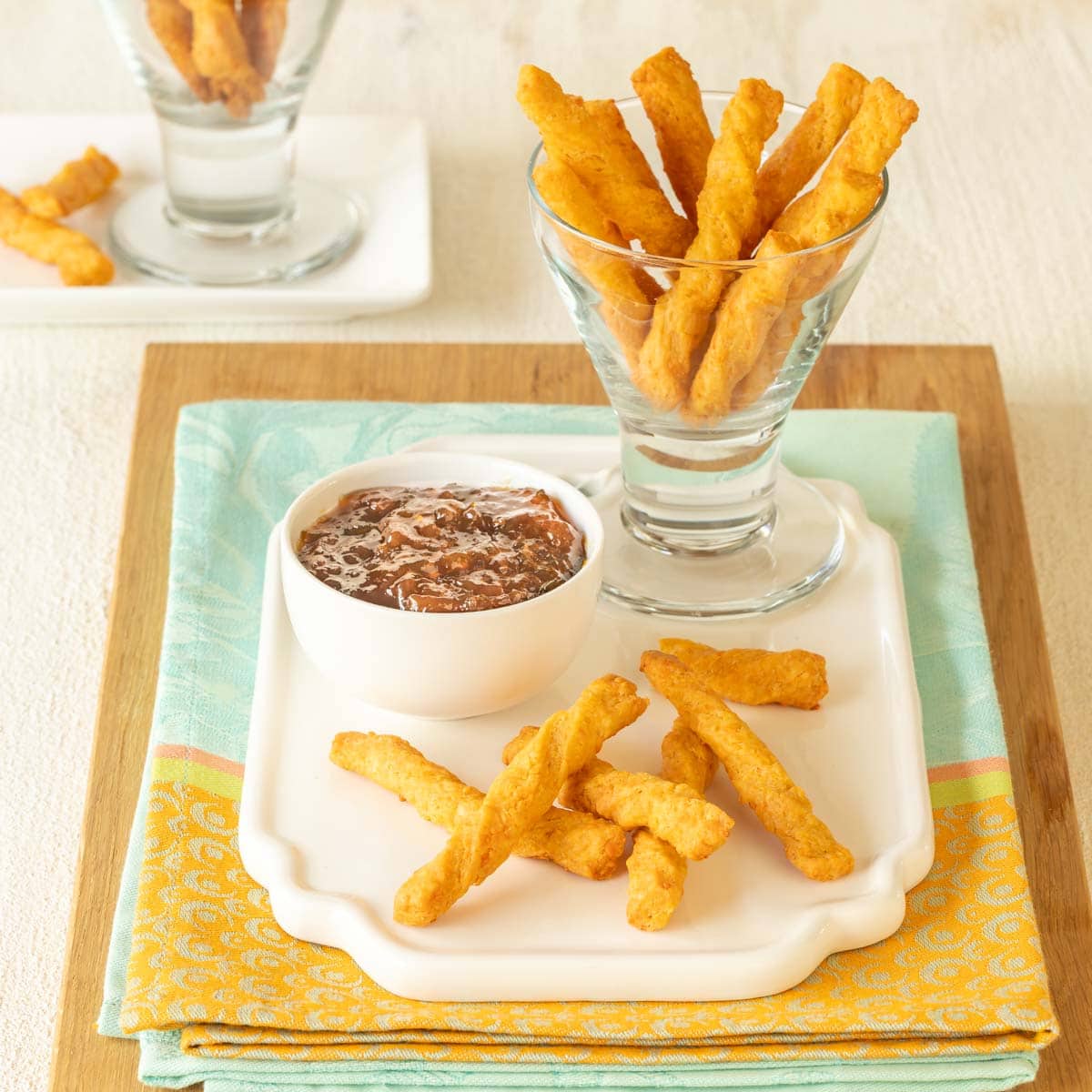 a glass holding cheese straws atop a white platter, with a few cheese straws in front and dipping sauce on the side