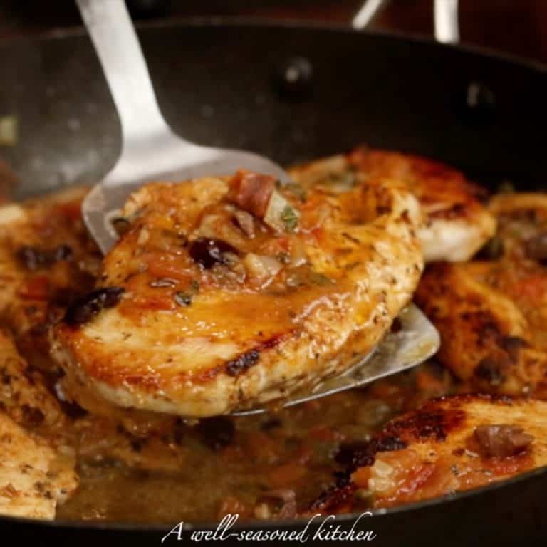 one cooked chicken breast on a metal spatula, over a pan with more chicken and. tomato-olive sauce