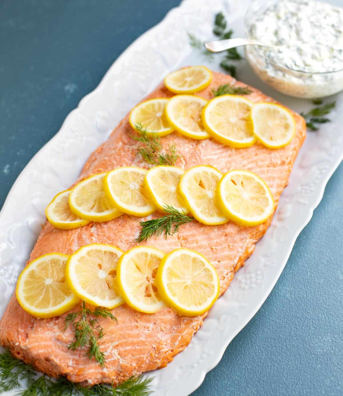 Oven poached salmon garnished with lemon slices and dill, with a side of cucumber dill sauce