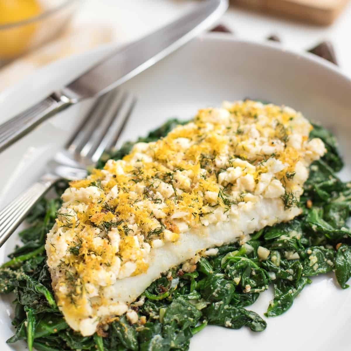 single serving of Baked Cod with Lemon, Feta & Dill on Spinach on a white plate with a fork & knife