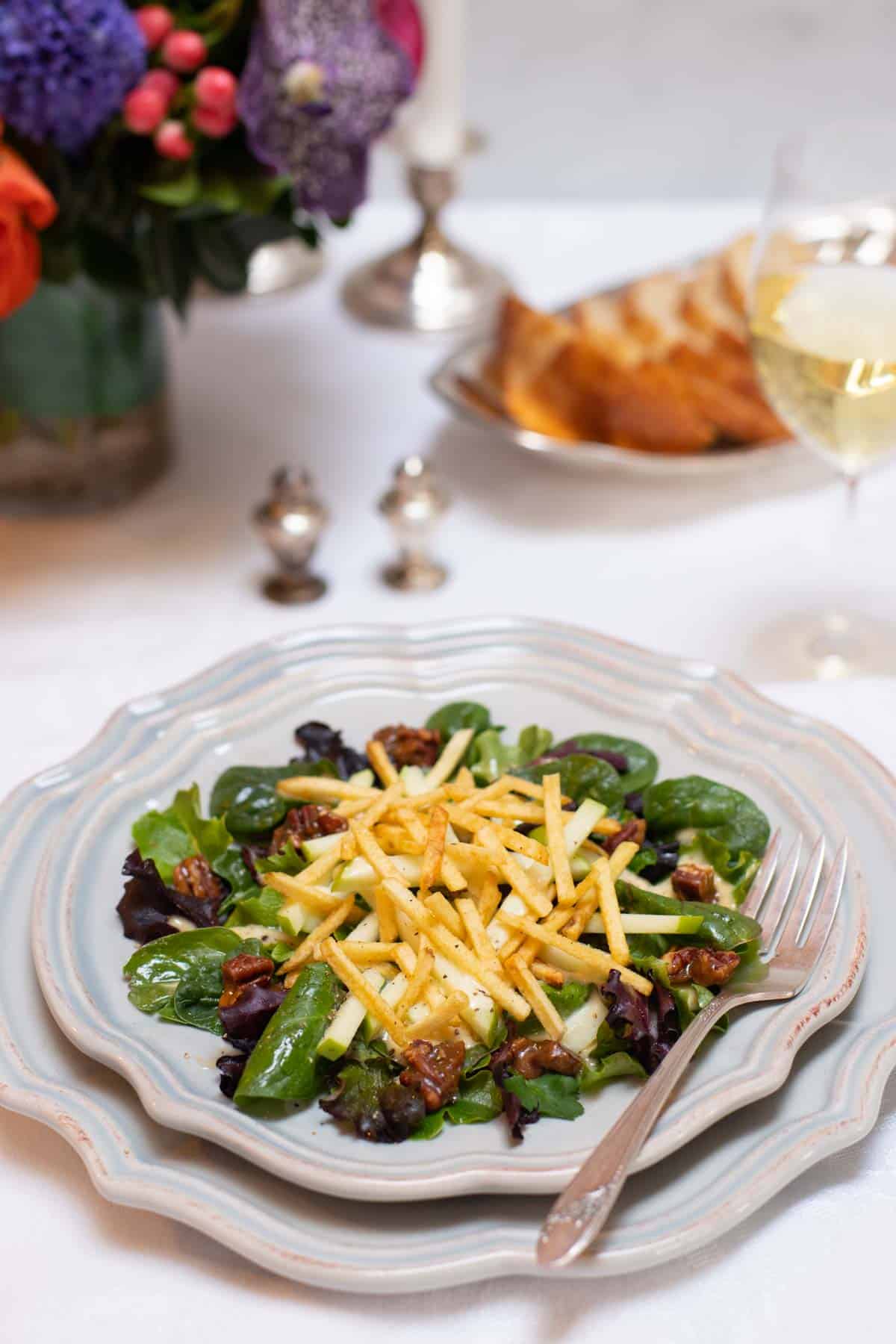 individual salad plate holding mixed greens sald with apples and pecans, on top of a dinner plate with bread and flowers in the background