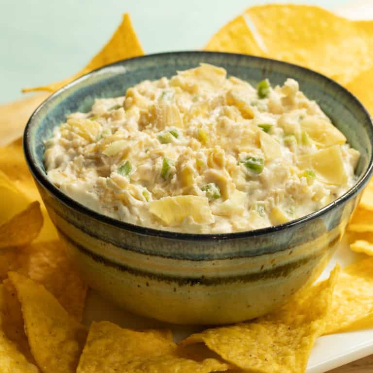 Blue bowl filled with Artichoke, Jalapeno and Cheese Dip, sitting on a white platter with tortilla chips