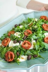 arugula spread on a blue platter, topped with roasted tomato halves, cheese and olives