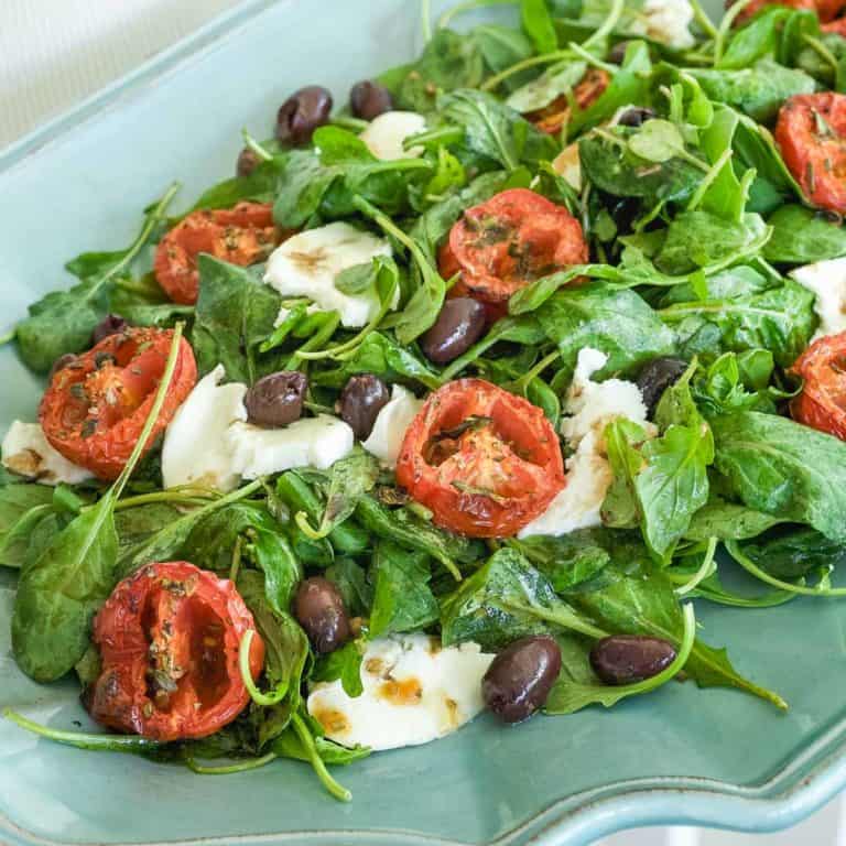 arugula spread on a blue platter, topped with roasted tomato halves, cheese and olives