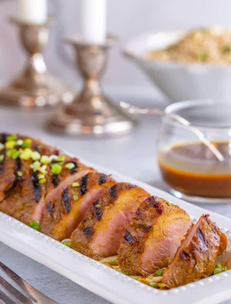 white platter filled with slices of Asian Pork Tenderloin. Bowl if rice and candles in the background