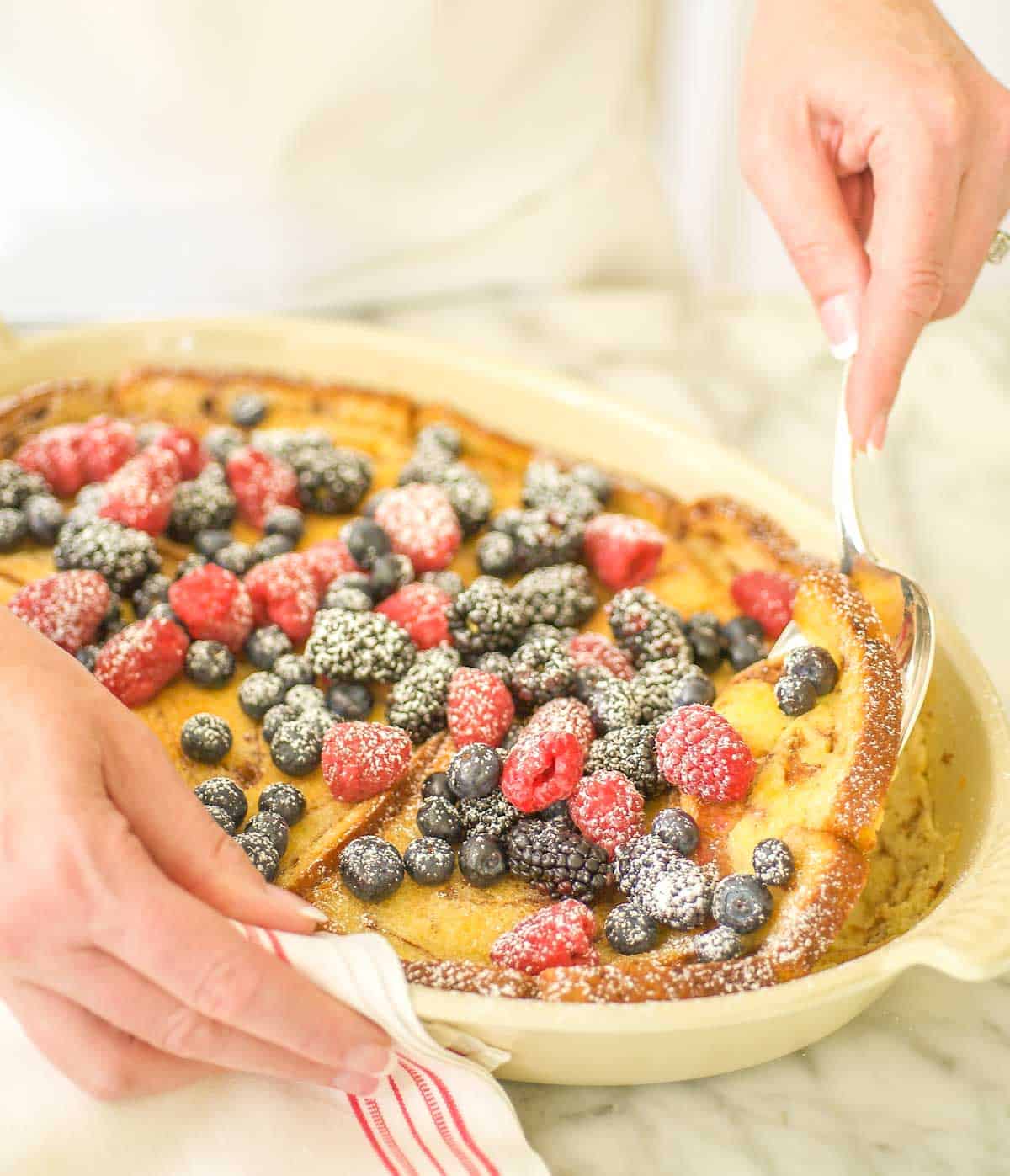 Oval baking dish filled with Baked Cinnamon French Toast, topped with berries and a spoon dishing out one serving.