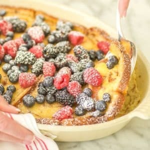 Oval baking dish filled with Baked Cinnamon French Toast, topped with berries and a spoon dishing out one serving