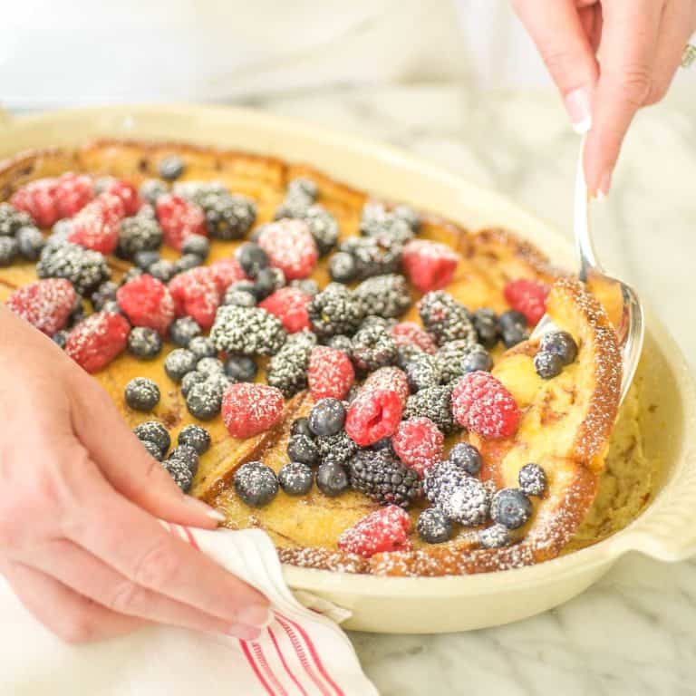 Oval baking dish filled with Baked Cinnamon French Toast, topped with berries and a spoon dishing out one serving