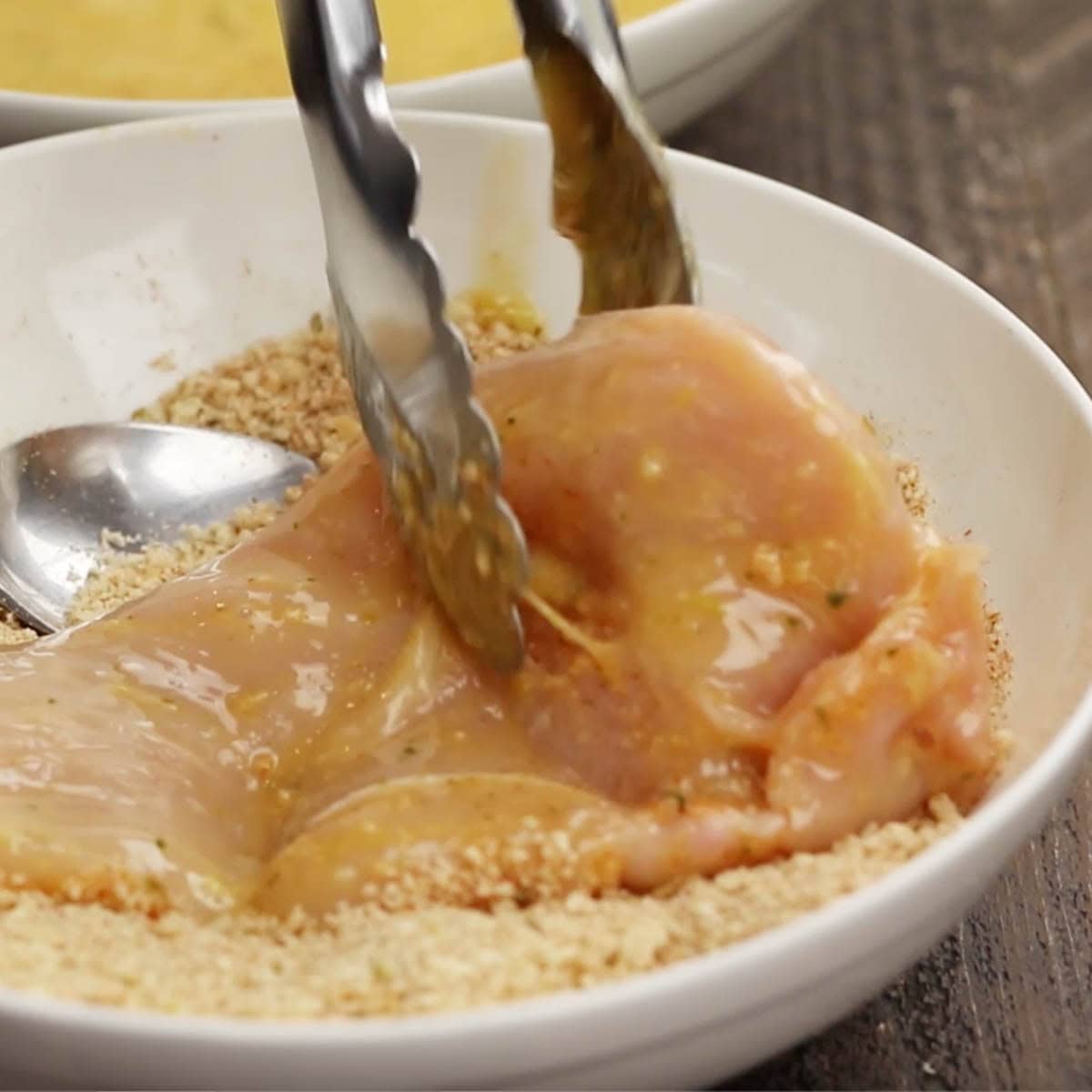 Tongs holding a chicken breast to dip in breading. 
