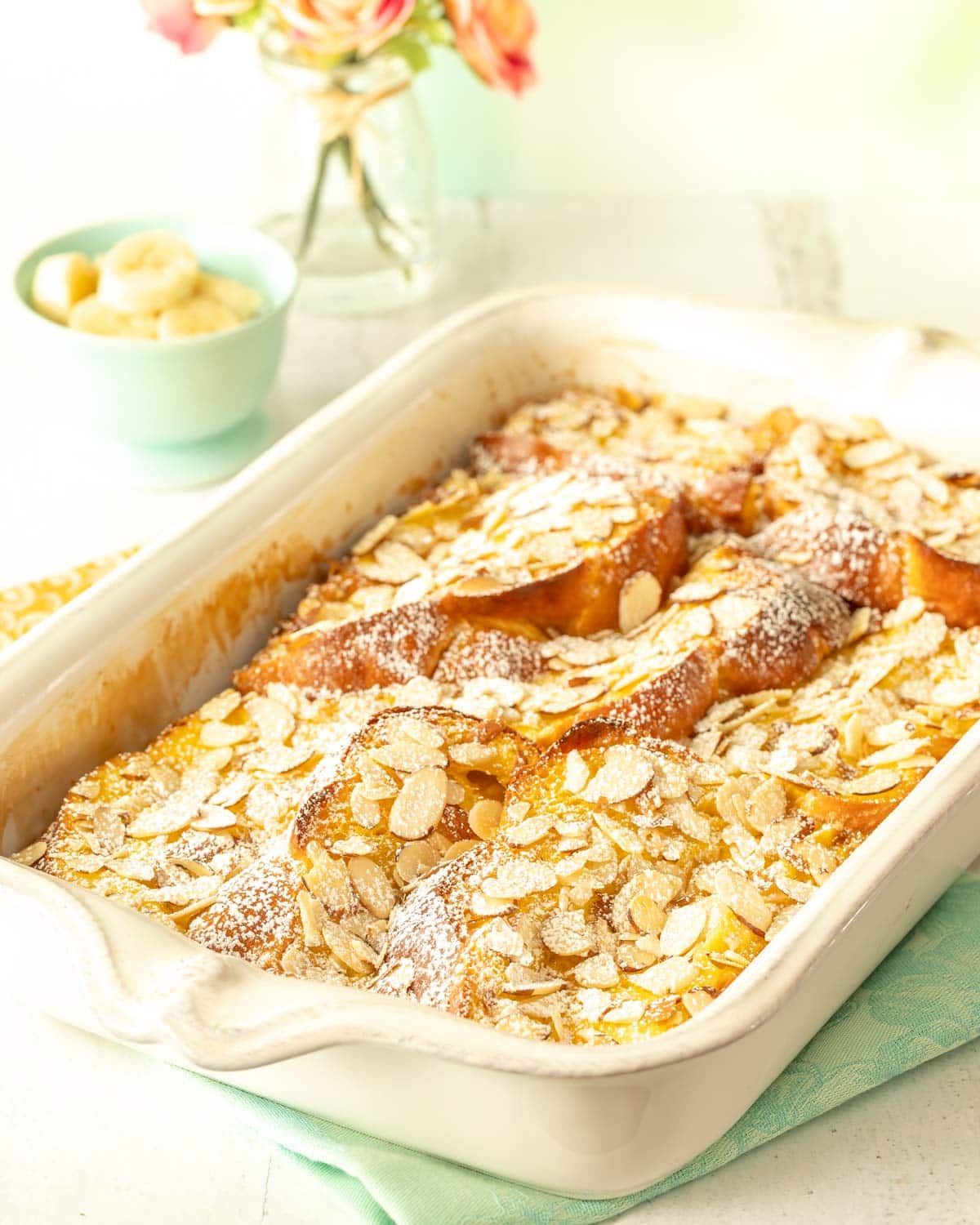 white casserole dish holding Baked Banana French Toast, with a small dish of bananas and flowers in back