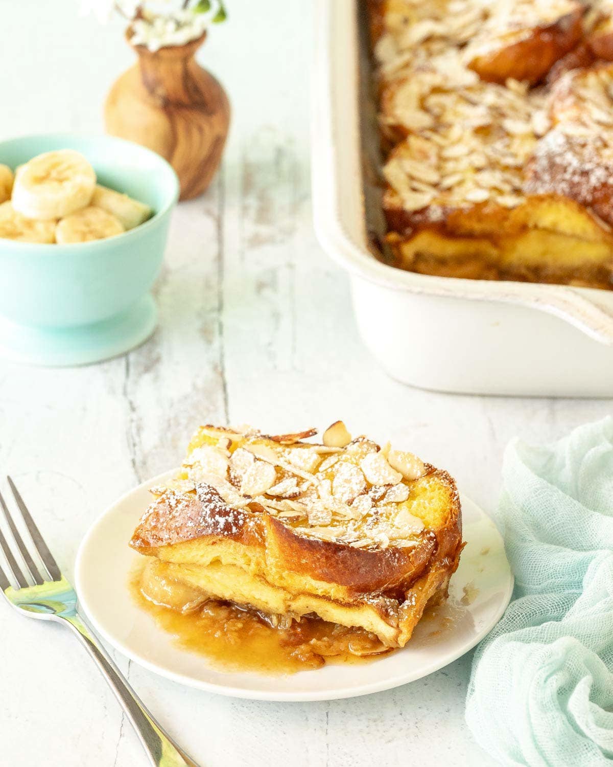Slice of Baked French Toast on a white plate with rest of casserole in the background alongside bowl of sliced bananas and flowers