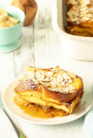 Slice of Baked French Toast on a white plate with rest of casserole in the background alongside bowl of sliced bananas and flowers