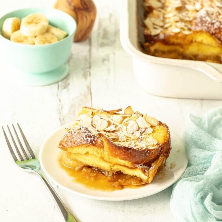 Slice of Baked French Toast on a white plate with rest of casserole in the background alongside bowl of sliced bananas and flowers