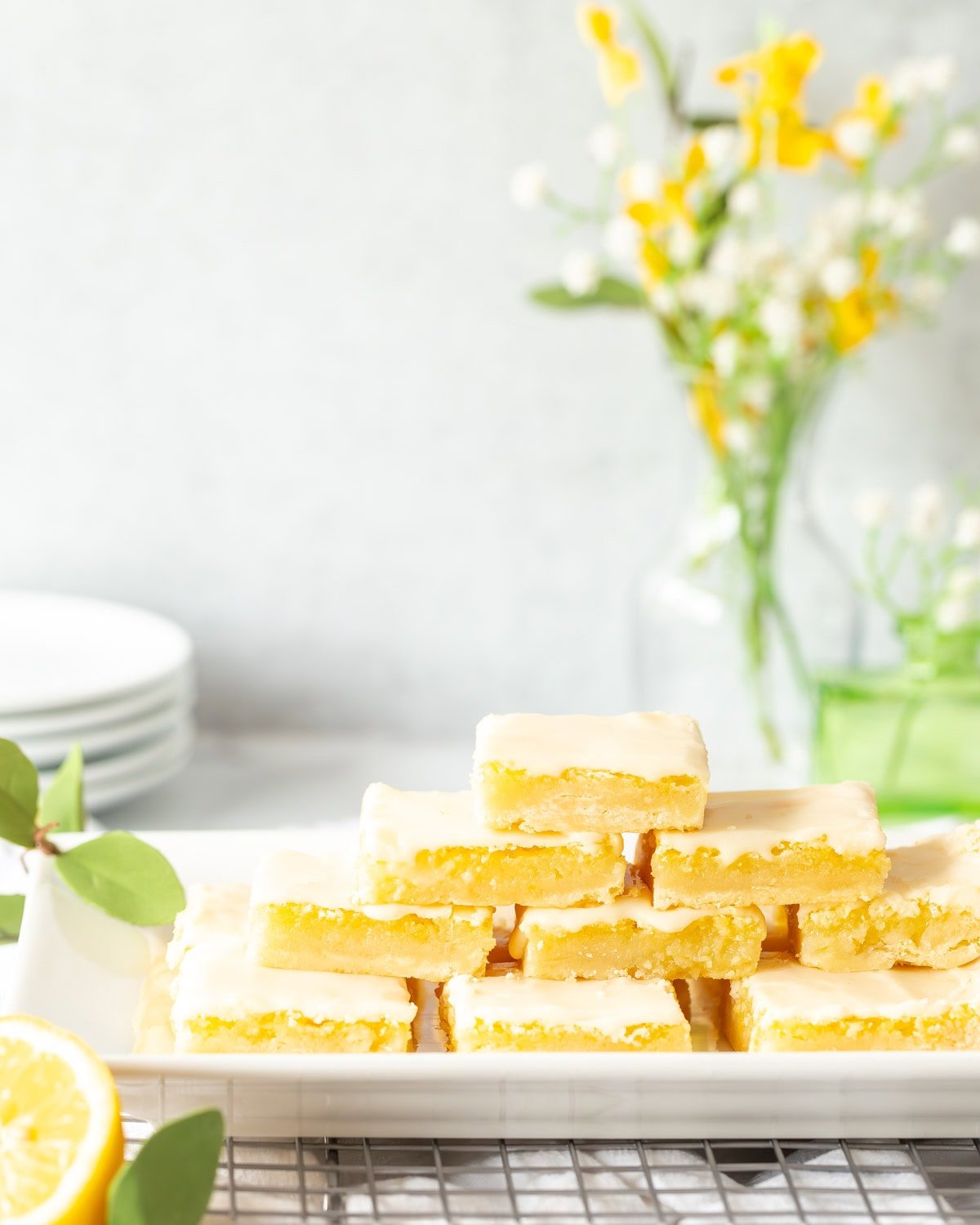 Homemade Lemon Bars stacked on a serving tray with small plates and flowers in the background