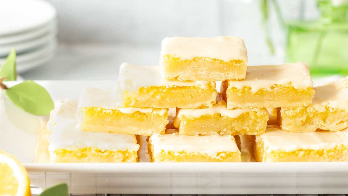 A stack of lemon bars topped with icing on a white platter, surrounded by green leaves and lemon slices.