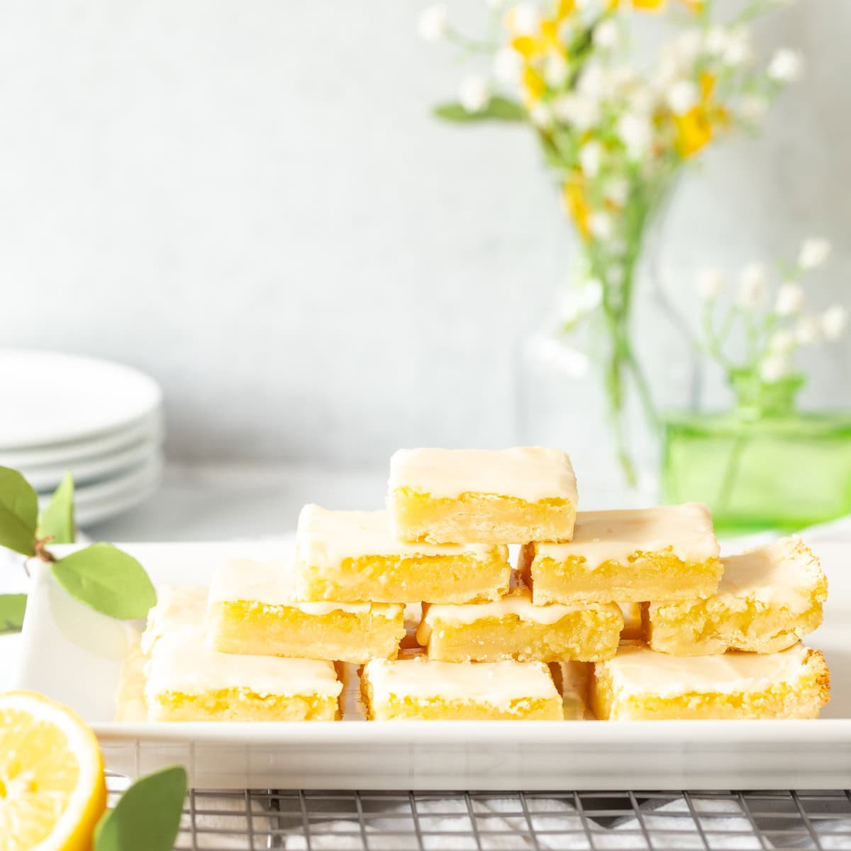 Homemade Lemon Bars stacked on a serving tray with small plates and flowers in the background