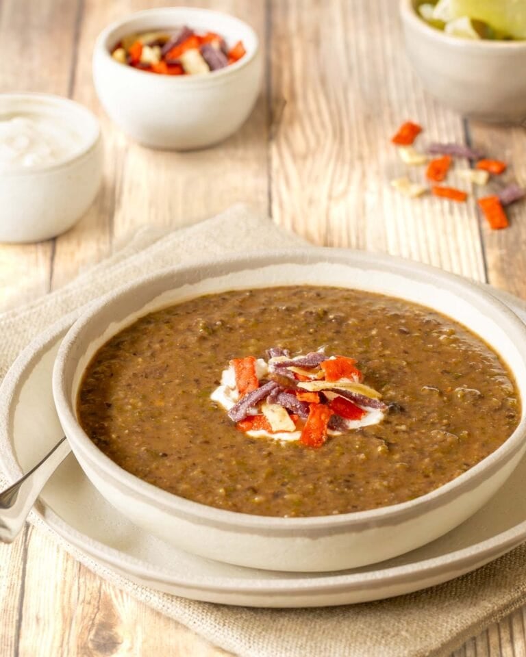 One serving of black bean soup garnished with sour cream and tortilla strips, with lime wedges, more sour cream and more strips in bowls on the side