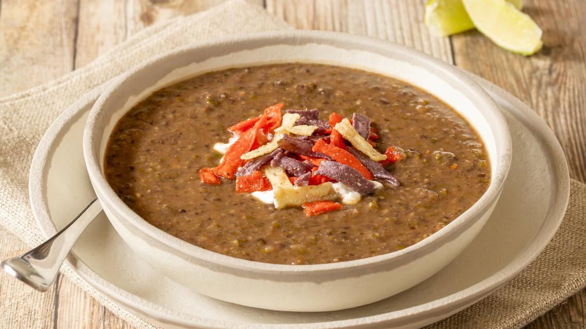 close up view of one serving of black bean soup garnished with sour cream and tortilla strips, with lime wedges on the side