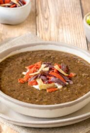 One serving of black bean soup garnished with sour cream and tortilla strips, with lime wedges, more sour cream and more strips in bowls on the side