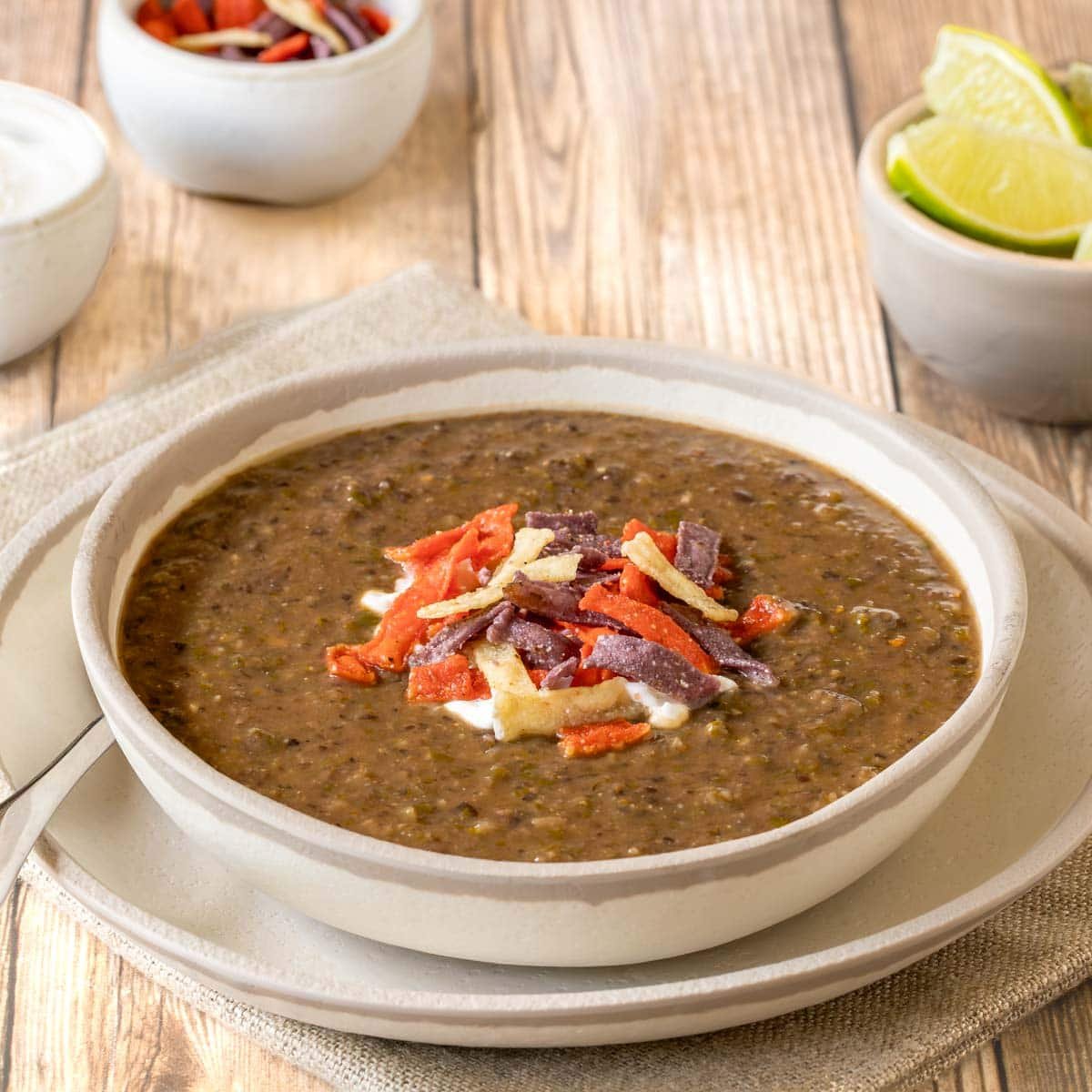 One serving of black bean soup garnished with sour cream and tortilla strips, with lime wedges, more sour cream and more strips in bowls on the side