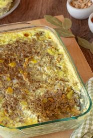 Bobotie with a corner removed, showing the inside. Removed piece is on a plate in the backgrounds, with a small dish of slivered almonds and another holding apricot jam