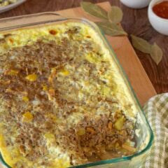 Bobotie with a corner removed, showing the inside. Removed piece is on a plate in the backgrounds, with a small dish of slivered almonds and another holding apricot jam