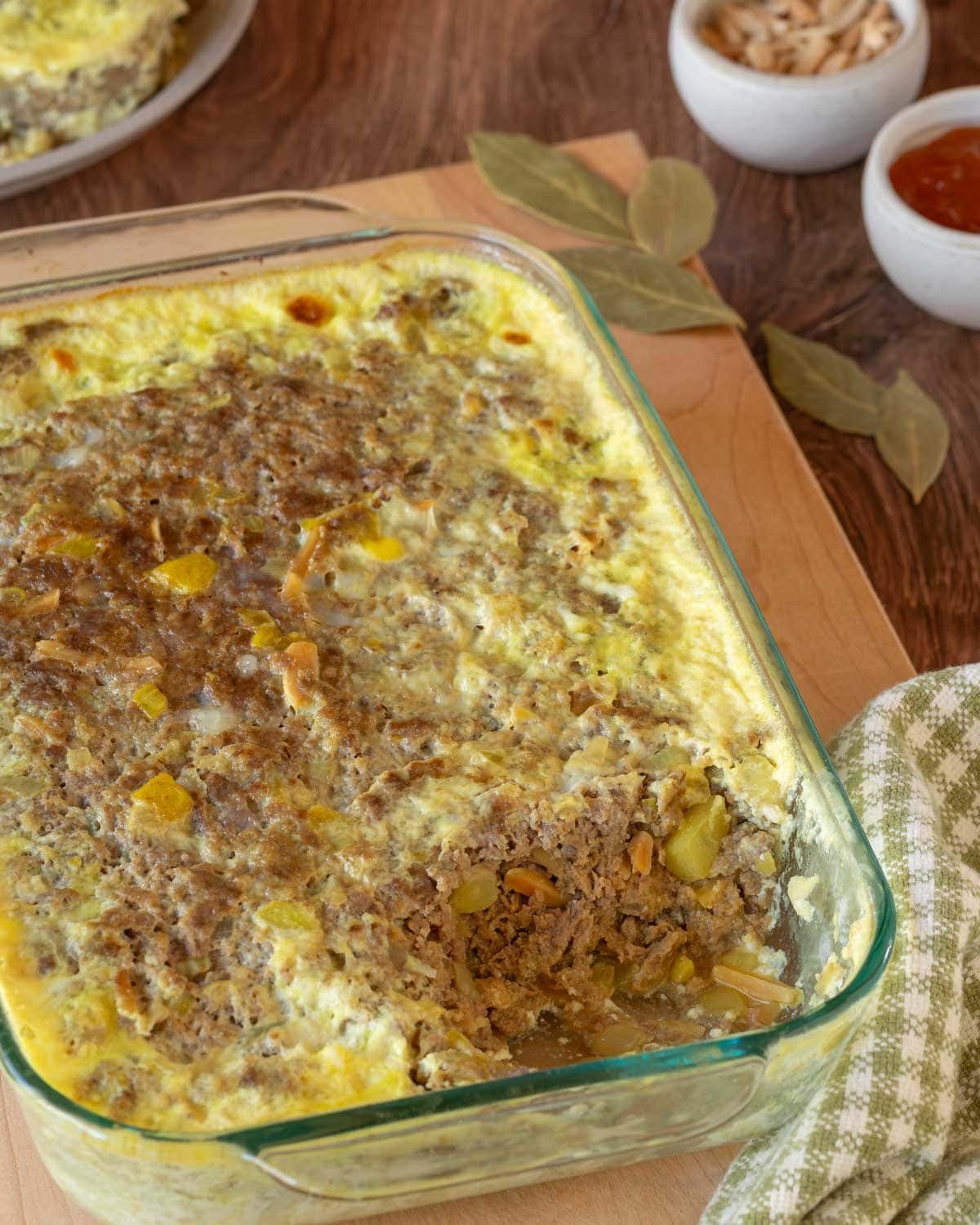 Bobotie with a corner removed, showing the inside. Removed piece is on a plate in the backgrounds, with a small dish of slivered almonds and another holding apricot jam