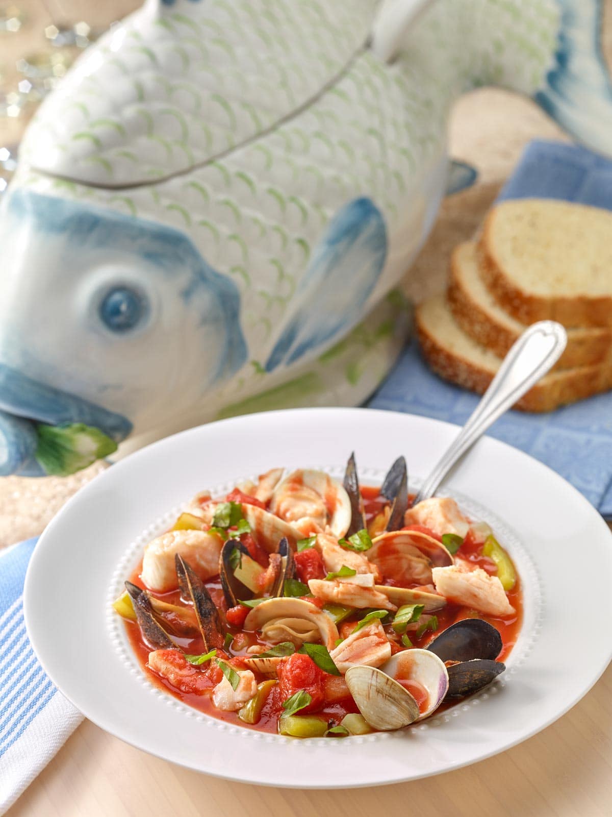 White bowl holding a portion of Bouillabaisse with a fish-shaped soup tureen and sliced bread in the background