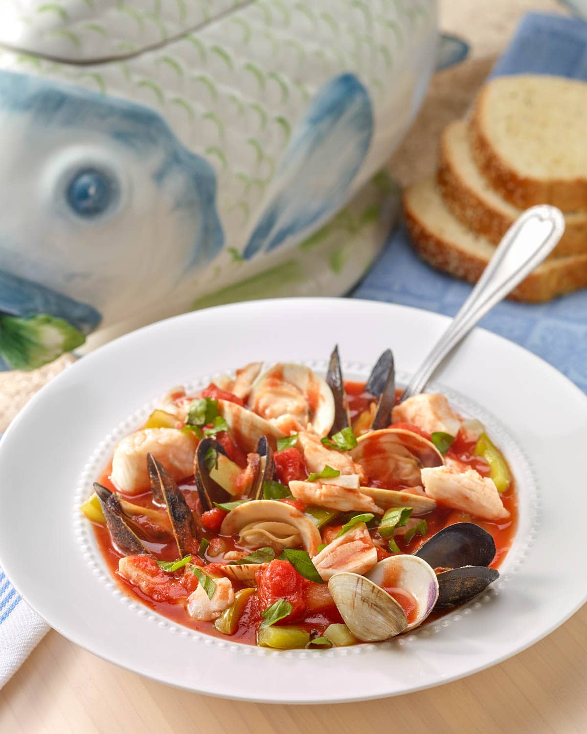 White bowl holding a portion of Bouillabaisse with a fish-shaped soup tureen and sliced bread in the background