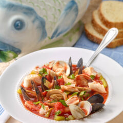 White bowl holding a portion of Bouillabaisse with a fish-shaped soup tureen and sliced bread in the background