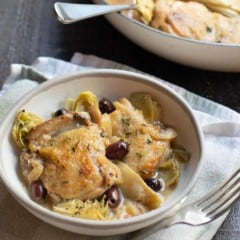 two braised chicken thighs with olives and artichoke hearts in a shallow bowl