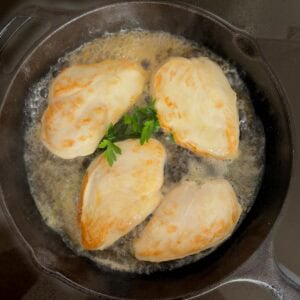 Cast iron skillet showing browned chicken breasts in skillet after liquid and seasonings have been added.