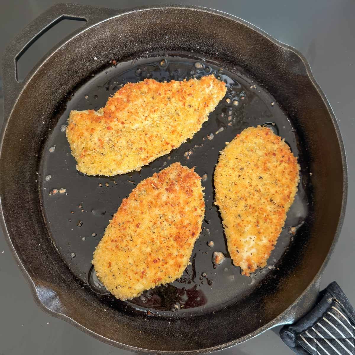 three breaded chicken breasts searing in a large skillet, showing light brown coating.