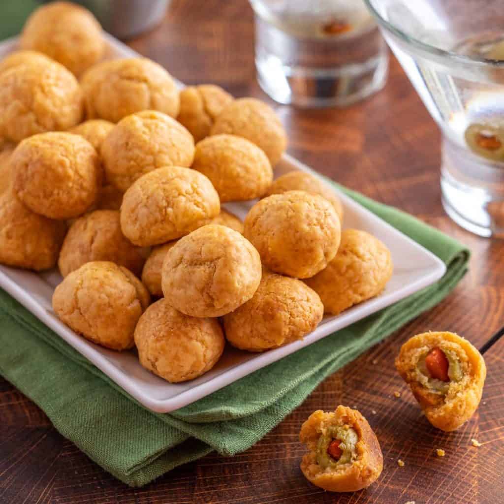 White rectangular platter filled with Olive Cheese Balls. In front are two cheese balls cut in half, to show olive inside