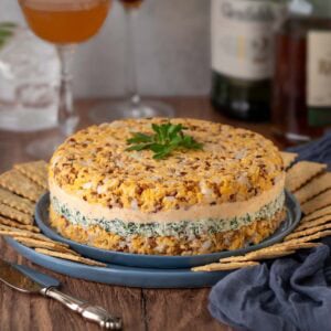 Closer view of Cheddar Cheese Torte on a blue plate surrounded by crackers, with bottles of alcohol and 3 mixed cocktails in the background