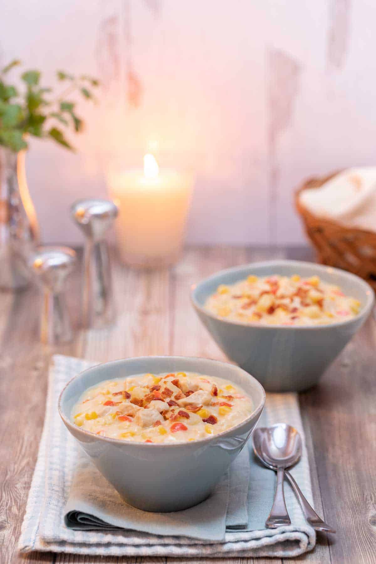 two bowls filled with Corn and Chicken Chowder with a basket of bread and a candle in the background.