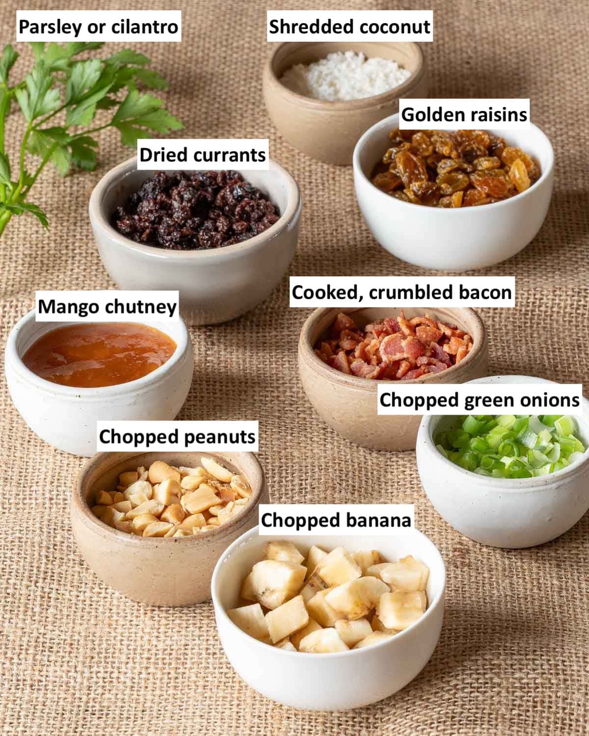 Condiments for boneless chicken curry including bacon, peanuts, chutney, raisins, currants, coconut, banana, green onion, and fresh herbs.