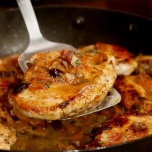 one cooked chicken breast on a metal spatula, over a pan with more chicken and tomato-olive sauce