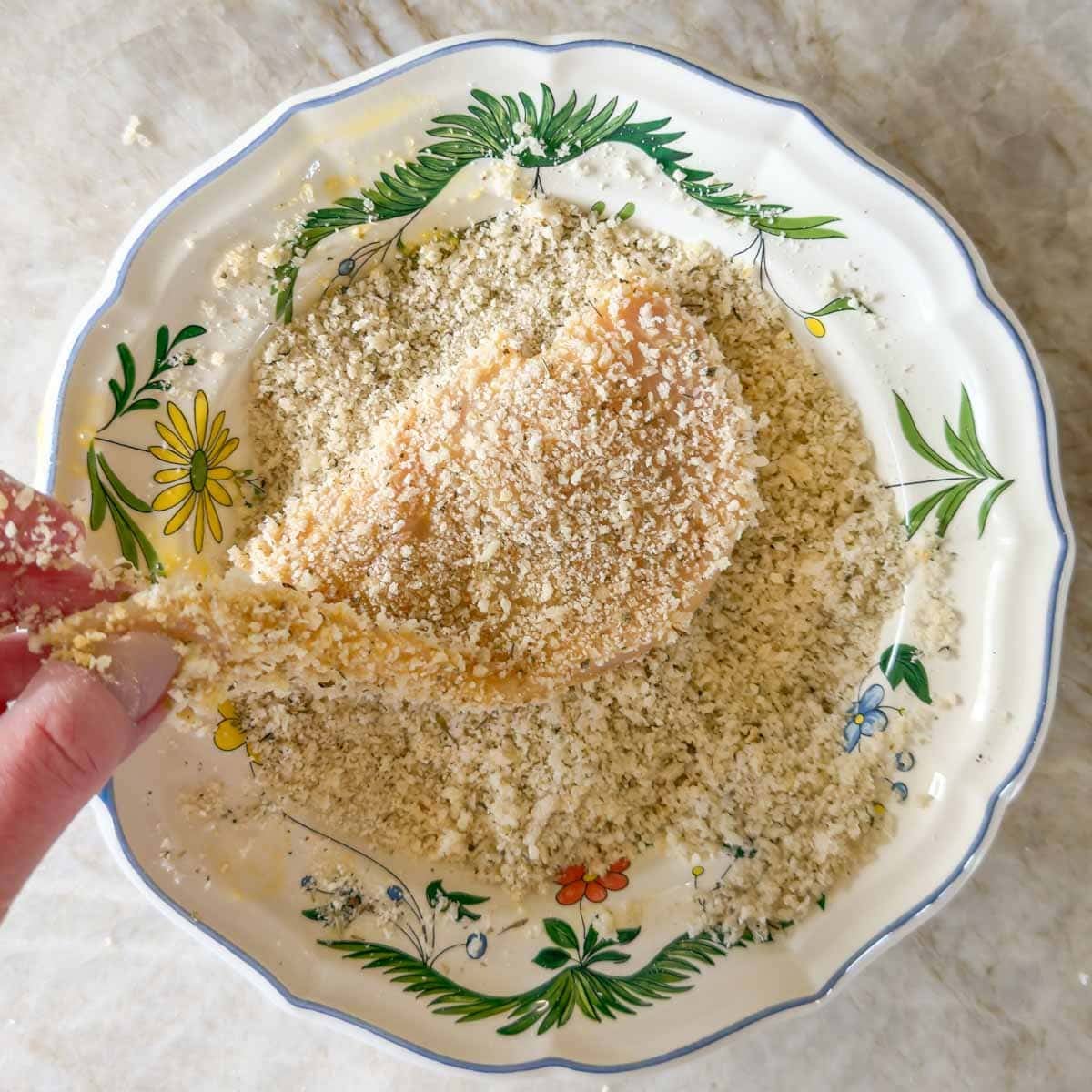 Single chicken breast being dipped in seasoned breadcrumbs, the last step in breading for chicken parmesan.