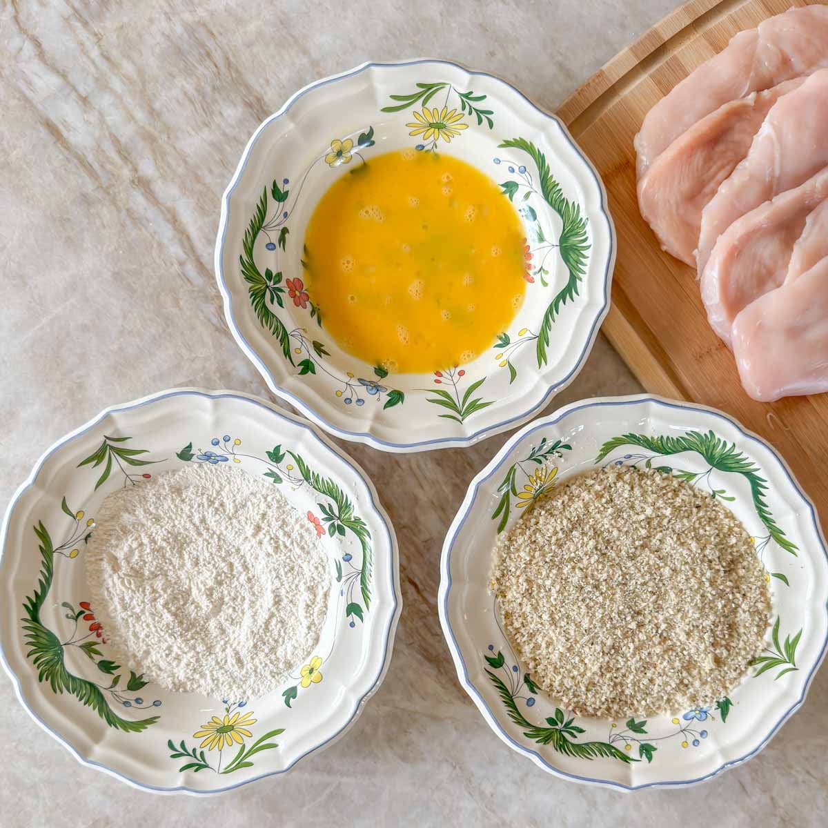 Three shallow bowls holding beaten egg, seasoned flour, and saasoned breadcrumbs, ready for dredging chicken.