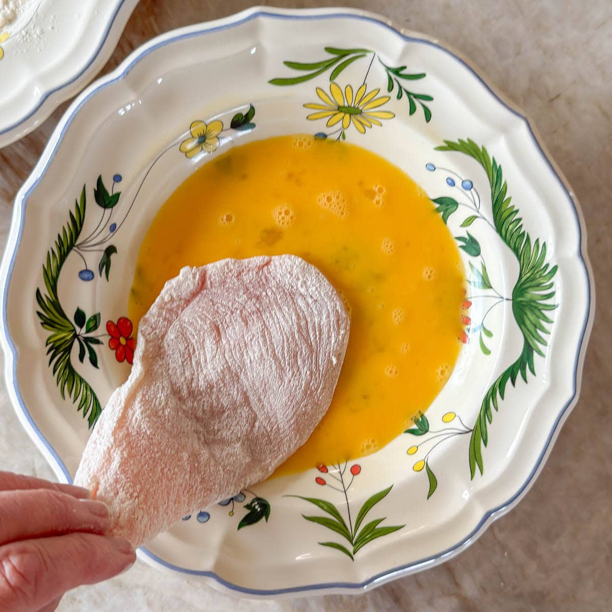 Single chicken breast already floured, being dipped in beaten egg, the 2nd step in breading for chicken parmesan.