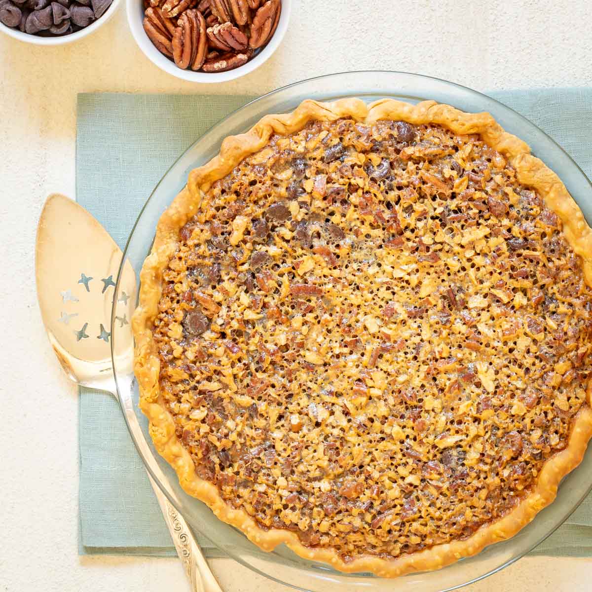 Overhead shot of Chocolate Pecan Pie, with a server and 2 bowls of pecans and chocolate chips on the side