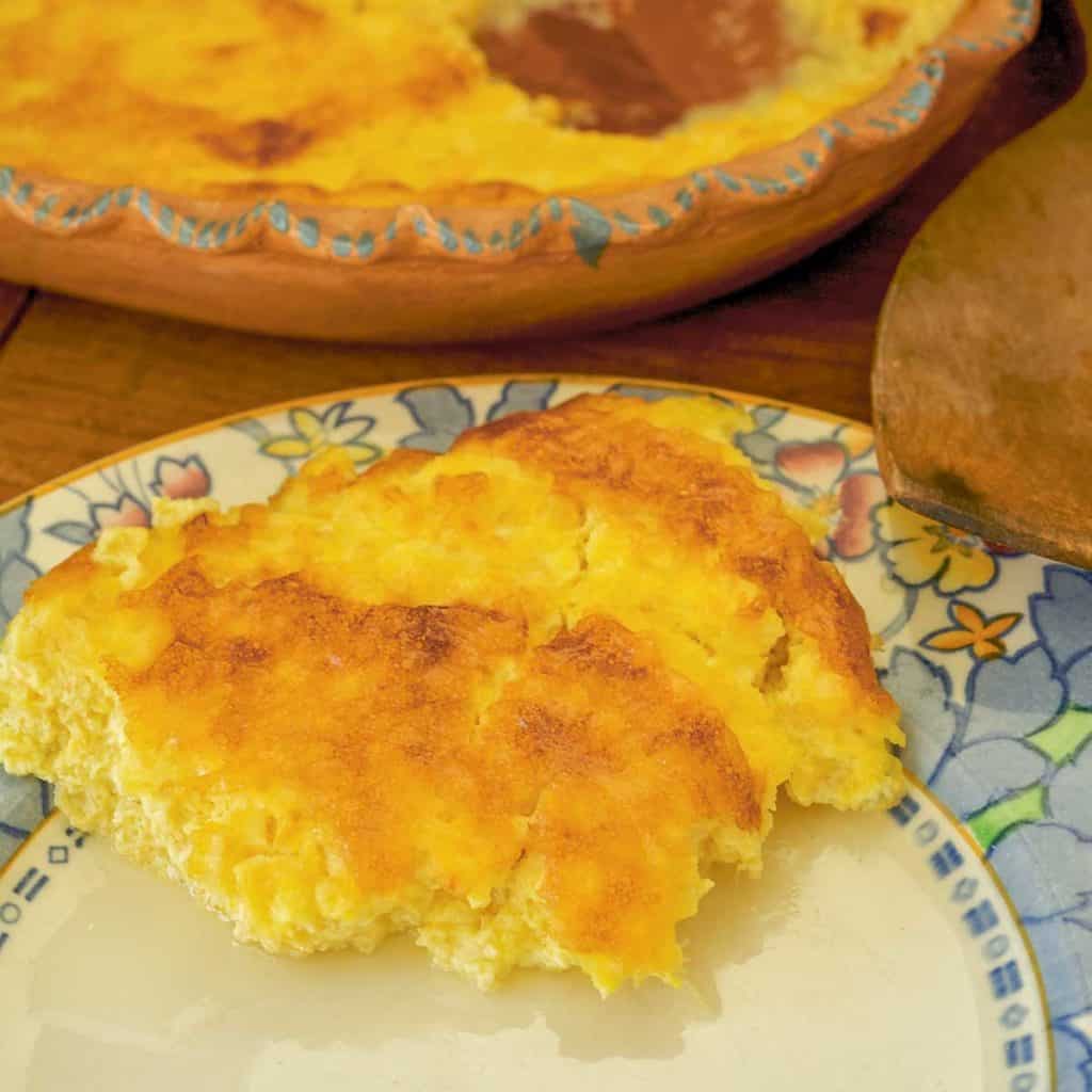 Plate showing a single serving of Corn Pudding with part of remainder of dish in the background