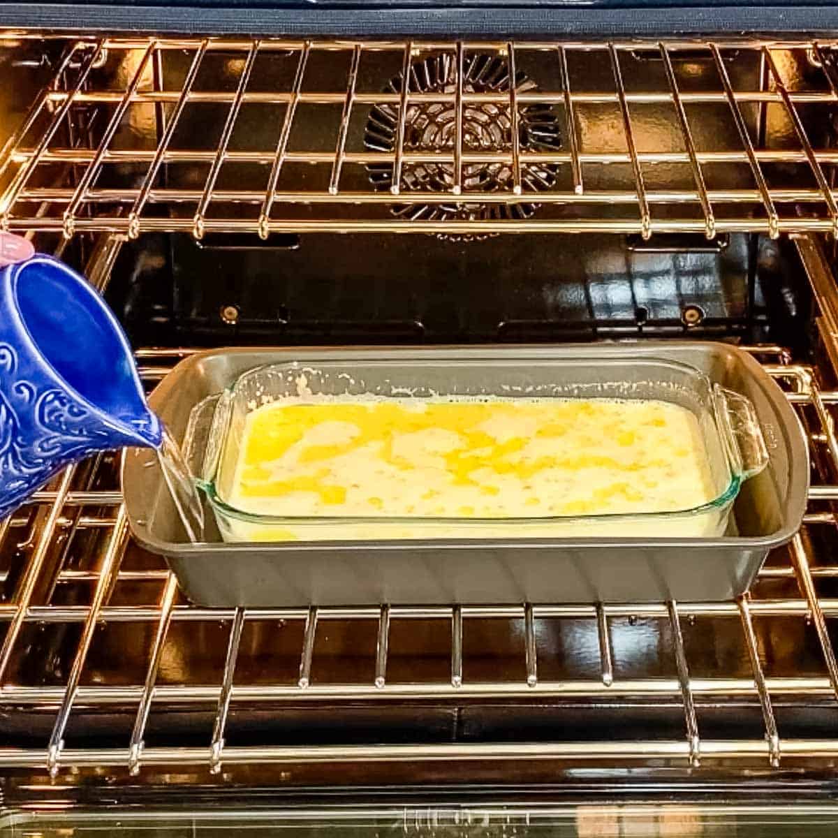 Corn pudding inside baking dish in oven with person pouring water into second outer baking dish